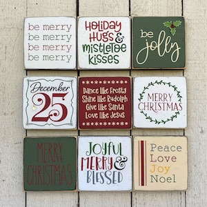 May include: Nine square wooden blocks with Christmas-themed text in various colors. Phrases include "be merry," "Holiday Hugs & mistletoe kisses," "be jolly," "December 25," "Merry Christmas," and "Peace Love Joy Noel."