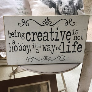 May include: White wooden sign with black text that reads "being creative is not a hobby, it's a way of life". The sign has decorative swirls at the top and bottom.