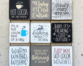 Könnte beinhalten: Neun quadratische Holzschilder mit winterlichen Phrasen und Grafiken. Die Schilder zeigen Texte wie "Hot Cocoa", "Baby It's Cold Outside" und "Fuzzy Socks". Farben sind Schwarz, Weiß, Grau und Rot.