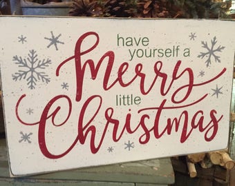 May include: A rectangular wooden sign with a white background and distressed edges. The sign features the phrase "have yourself a Merry little Christmas" in red script, with green and gray accents. Snowflakes are scattered around the text.