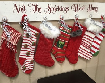 Op de afbeelding: Een witte houten kousenhouder met de tekst "And The Stockings Were Hung" in rode cursieve letters. Zeven rood-wit gestreepte kousen hangen aan de houder, elk met een wit naamschildje. De namen op de kousen zijn:  "Jamition", "Madison", "Lexie", "Millie", "Gavin", "Gunner" en "Nora".