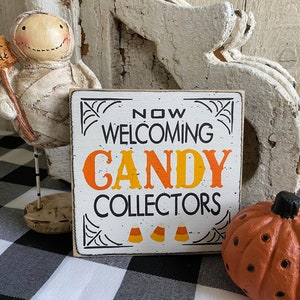 May include: A Halloween-themed display featuring a mummy figurine holding a "Trick or Treat" sign, a sign that reads "Now Welcoming Candy Collectors", and a small orange pumpkin. The sign has a white background with black and orange text.