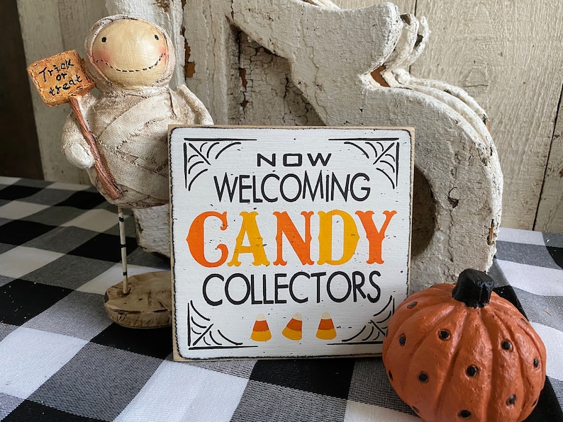 May include: A Halloween-themed display featuring a mummy figurine holding a "Trick or Treat" sign, a sign that reads "Now Welcoming Candy Collectors", and a small orange pumpkin. The sign has a white background with black and orange text.