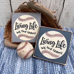 Op de afbeelding: Twee houten borden met een baseballthema. De borden zijn blauw en wit geschilderd en hebben een baseball met de tekst "Living Life by the seams".