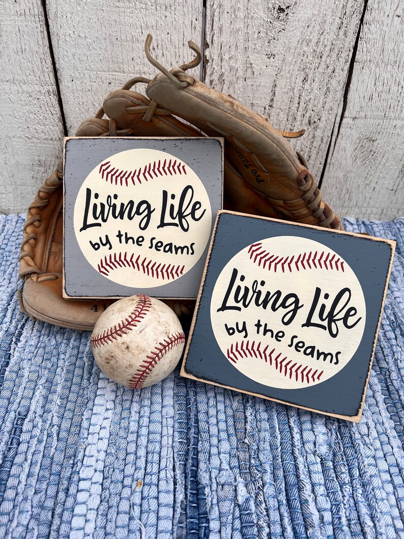 Op de afbeelding: Twee houten borden met een baseballthema. De borden zijn blauw en wit geschilderd en hebben een baseball met de tekst "Living Life by the seams".