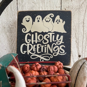 May include: A black wooden sign with the words "Ghostly Greetings" and four white ghost figures. The sign is set against a rustic white wood background. A green metal truck bed filled with orange pumpkins is in the foreground.