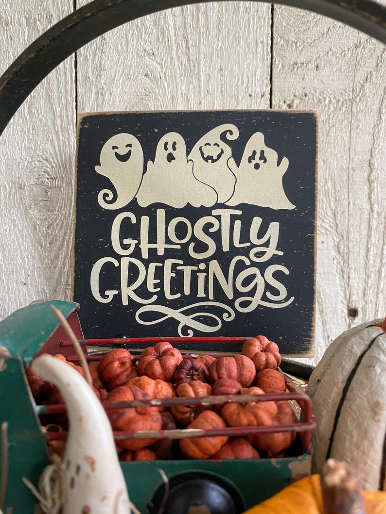 May include: A black wooden sign with the words "Ghostly Greetings" and four white ghost figures. The sign is set against a rustic white wood background. A green metal truck bed filled with orange pumpkins is in the foreground.