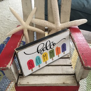 May include: A white wooden chair with red accents and a sign that says "hello summer" with colorful popsicle graphics. Two white starfish are on the chair.