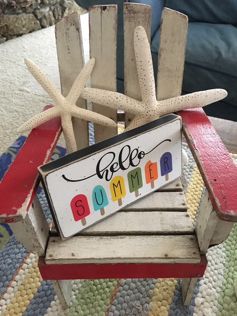 May include: A white wooden chair with red accents and a sign that says "hello summer" with colorful popsicle graphics. Two white starfish are on the chair.