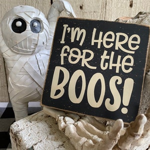 May include: A Halloween-themed decorative sign with the text "I'm HERE for the BOOS!" in cream lettering on a black background. A white mummy figure and a skeleton hand are in the background, creating a spooky atmosphere.