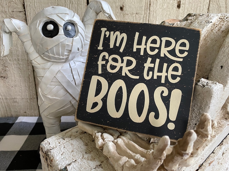 May include: A Halloween-themed decorative sign with the text "I'm HERE for the BOOS!" in cream lettering on a black background. A white mummy figure and a skeleton hand are in the background, creating a spooky atmosphere.