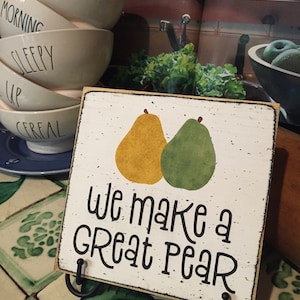 May include: A wooden sign with a white background and black text that reads "We make a great pear". Two pears are painted on the sign, one yellow and one green. The sign is on a black stand.
