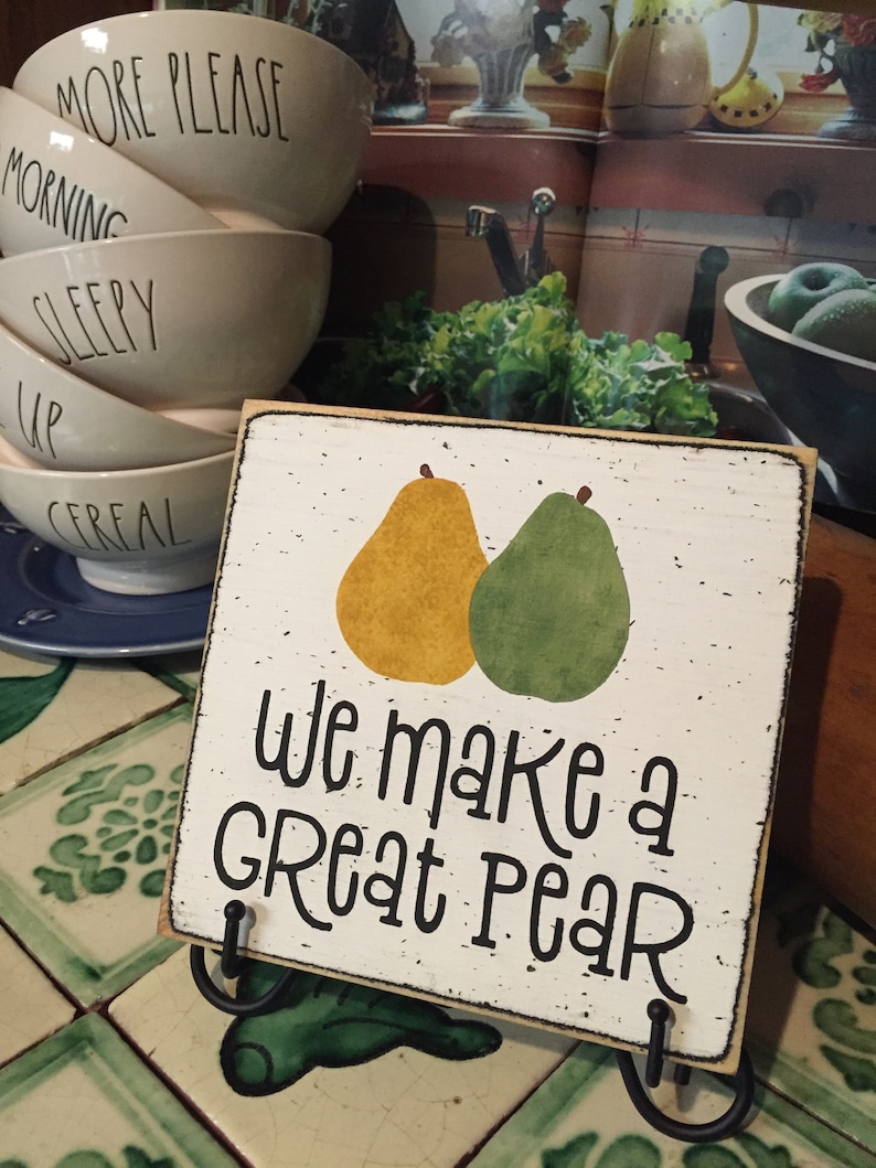 May include: A wooden sign with a white background and black text that reads "We make a great pear". Two pears are painted on the sign, one yellow and one green. The sign is on a black stand.