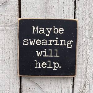 May include: Black wooden sign with white text that reads "Maybe swearing will help."