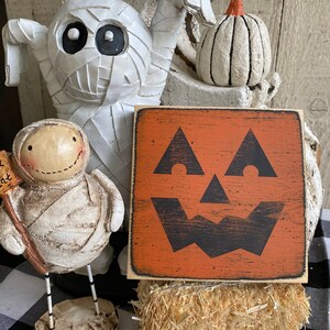 May include: Halloween-themed decorations featuring a mummy figure, a small pumpkin, and a wooden sign with a jack-o'-lantern face. The sign is orange with a black face, and the decorations are set on a hay bale.