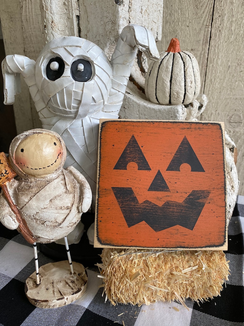 May include: Halloween-themed decorations featuring a mummy figure, a small pumpkin, and a wooden sign with a jack-o'-lantern face. The sign is orange with a black face, and the decorations are set on a hay bale.