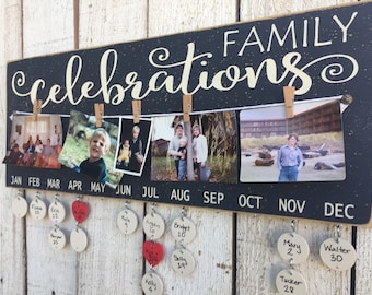 May include: A rectangular black wooden sign with the text FAMILY celebrations in white script. A white string holds five photographs with wooden clips. Below, the months JAN through DEC are printed with hanging white and red circular tags.