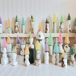 May include: A collection of colorful bottle brush trees in various sizes and pastel hues, displayed on distressed white wooden pedestals and bases. The trees are arranged on a white shelf and tabletop against a white shiplap wall, creating a whimsical, vintage aesthetic.