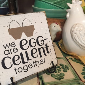 May include: White wooden sign with black text that reads "We are egg-cellent together". The sign has a brown silhouette of two eggs in a carton. The sign is sitting on a table with a white ceramic chicken figurine.