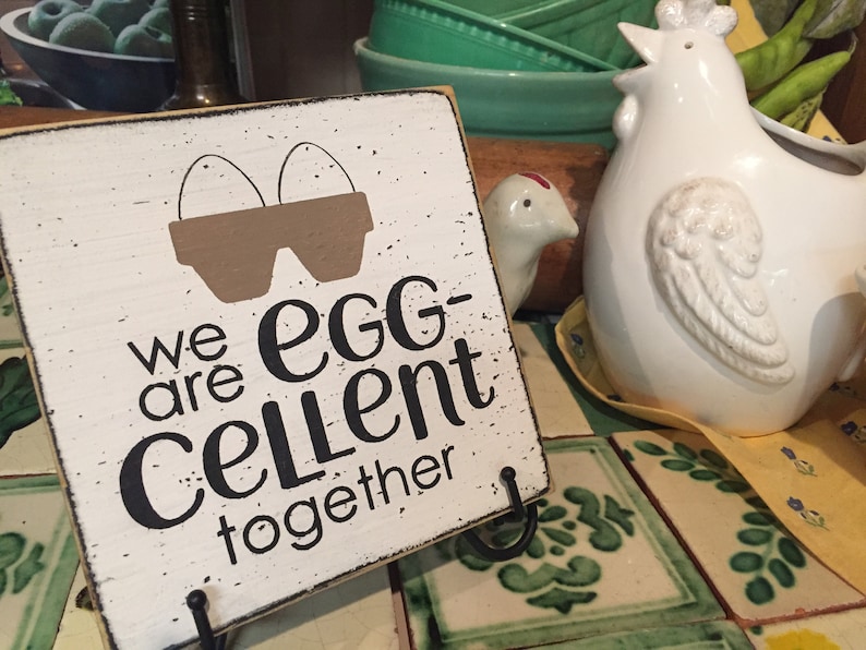 May include: White wooden sign with black text that reads "We are egg-cellent together". The sign has a brown silhouette of two eggs in a carton. The sign is sitting on a table with a white ceramic chicken figurine.