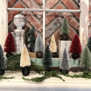 May include: A collection of miniature bottle brush Christmas trees in various colors, including green, red, and cream. The trees are arranged on a shelf in front of a brick wall and a distressed white window frame. The trees are approximately 6 inches tall.