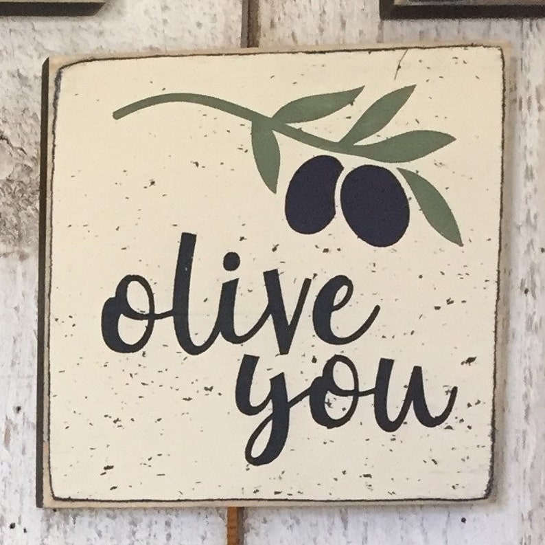 May include: A white wooden sign with black lettering that reads "olive you". The sign has a green olive branch with two black olives painted on it.
