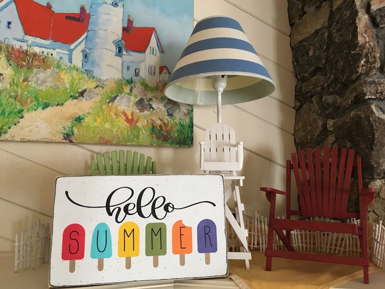 May include: A white wooden sign with the text "hello summer" in black cursive script. The word "summer" is spelled out with colorful popsicle graphics.