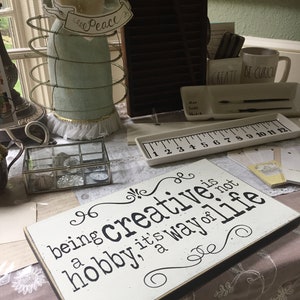 May include: A white wooden sign with black text that reads "Being creative is a hobby, it's a way of life". The sign is sitting on a table with other decorative items.