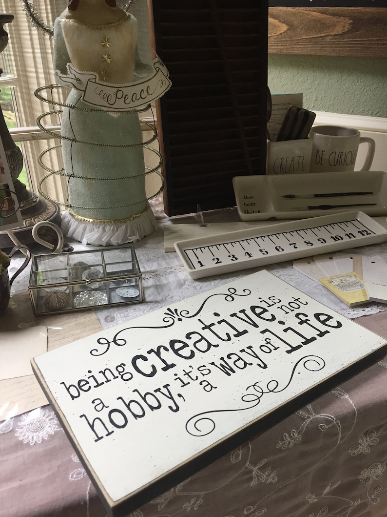 May include: A white wooden sign with black text that reads "Being creative is a hobby, it's a way of life". The sign is sitting on a table with other decorative items.