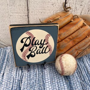 Op de afbeelding: Een houten bord met een baseball-afbeelding en de tekst "Play Ball" in zwarte schrijfletters. Het bord is geschilderd in tinten blauw en wit en heeft een verweerde uitstraling. Het bord staat op een blauw-wit geweven tapijt naast een baseball en een bruine leren honkbalhandschoen.