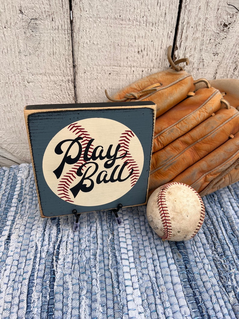 Op de afbeelding: Een houten bord met een baseball-afbeelding en de tekst "Play Ball" in zwarte schrijfletters. Het bord is geschilderd in tinten blauw en wit en heeft een verweerde uitstraling. Het bord staat op een blauw-wit geweven tapijt naast een baseball en een bruine leren honkbalhandschoen.