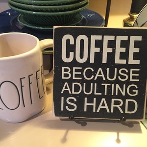 Peut inclure: Une tasse &agrave; caf&eacute; en c&eacute;ramique blanche avec le mot "COFFEE" &eacute;crit en noir sur le c&ocirc;t&eacute;, &agrave; c&ocirc;t&eacute; d'une pancarte en bois noir avec le texte "COFFEE BECAUSE ADULTING IS HARD".