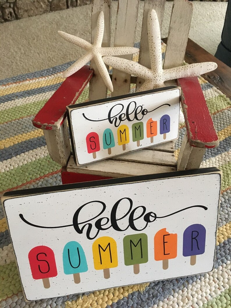 May include: Two wooden signs with the text "hello summer" in black cursive script. The signs feature a row of colorful popsicle graphics. The signs are sitting on a red and white striped beach chair.