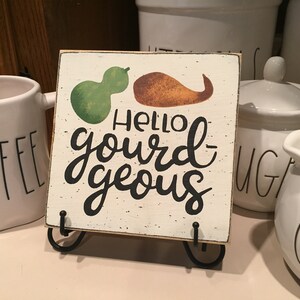 May include: A small wooden sign with a white background and black lettering that reads "Hello gourd-geous". The sign has a green and orange gourd painted on it and is sitting on a black metal stand.