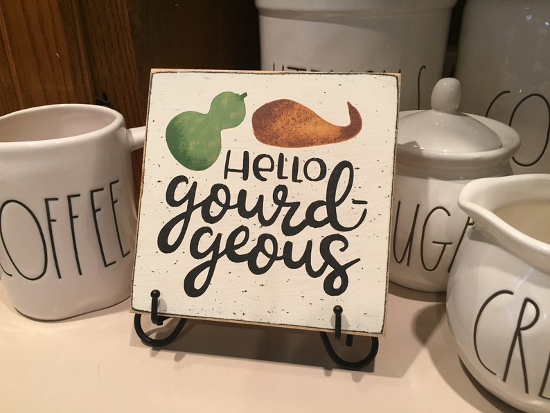 May include: A small wooden sign with a white background and black lettering that reads "Hello gourd-geous". The sign has a green and orange gourd painted on it and is sitting on a black metal stand.