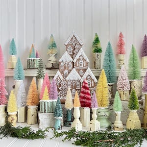 May include: A collection of colorful miniature Christmas trees and a gingerbread house. The trees are various shades of pink, green, yellow, and blue, and are mounted on decorative bases. The gingerbread house is decorated with white icing.