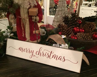 May include: A rectangular, off-white wooden sign with the words "merry christmas" in red script. The sign is part of a Christmas display, with a Santa Claus figure, pine cones, and red ornaments.