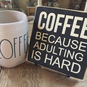 Peut inclure: Une tasse &agrave; caf&eacute; en c&eacute;ramique blanche avec le mot "COFFEE" imprim&eacute; en noir. Une pancarte en bois noir avec du texte blanc indique "COFFEE BECAUSE ADULTING IS HARD".
