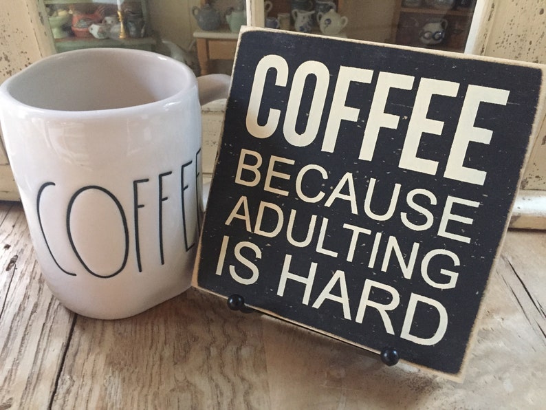 Peut inclure: Une tasse &agrave; caf&eacute; en c&eacute;ramique blanche avec le mot "COFFEE" imprim&eacute; en noir. Une pancarte en bois noir avec du texte blanc indique "COFFEE BECAUSE ADULTING IS HARD".