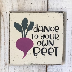 May include: A wooden sign with a distressed white background and black lettering. The sign features a cartoon beet with green leaves and a purple body. The text on the sign reads "Dance to your own beet".