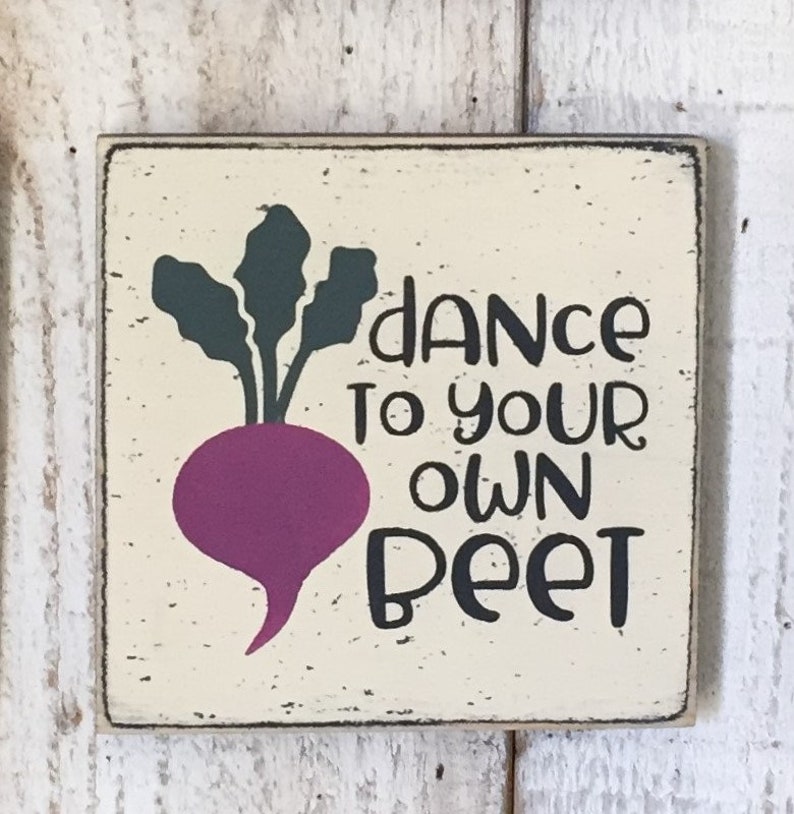 May include: A wooden sign with a distressed white background and black lettering. The sign features a cartoon beet with green leaves and a purple body. The text on the sign reads "Dance to your own beet".