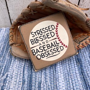 Op de afbeelding: Een houten bord met een witte cirkel en rode baseballnaden. Het bord staat "Stressed Blessed and Baseball Obsessed".