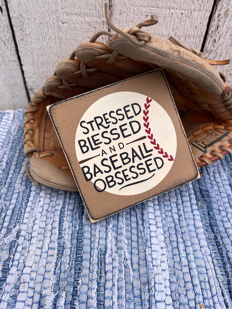 Op de afbeelding: Een houten bord met een witte cirkel en rode baseballnaden. Het bord staat "Stressed Blessed and Baseball Obsessed".