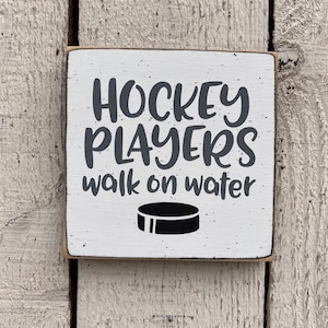 May include: White wooden sign with black text that reads "Hockey Players walk on water". A black hockey puck is below the text.