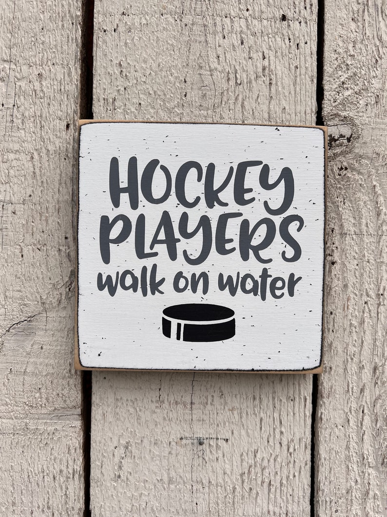 May include: White wooden sign with black text that reads "Hockey Players walk on water". A black hockey puck is below the text.