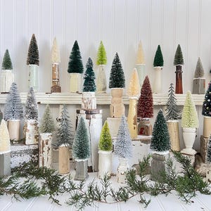 May include: A collection of decorative miniature Christmas trees in various colors and textures. The trees are displayed on white and distressed wooden pedestals, with greenery in the foreground. The trees vary in size and color, creating a festive display.