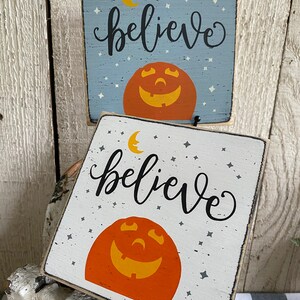 May include: Two square wooden signs with a Halloween theme. One is light blue, the other white. Both feature a smiling jack-o'-lantern, a crescent moon, stars, and the word "believe" in black script. The signs are rustic and decorative.