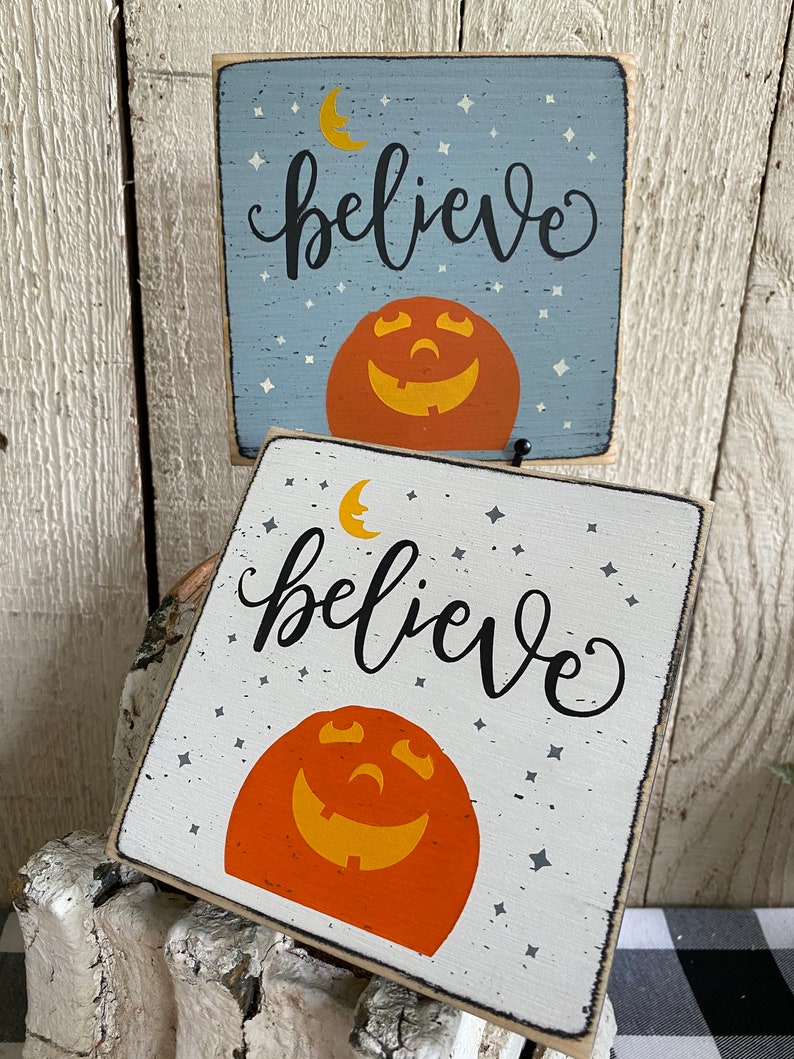 May include: Two square wooden signs with a Halloween theme. One is light blue, the other white. Both feature a smiling jack-o'-lantern, a crescent moon, stars, and the word "believe" in black script. The signs are rustic and decorative.
