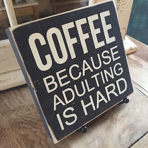 Peut inclure: Une pancarte en bois noir avec du texte blanc qui dit "COFFEE BECAUSE ADULTING IS HARD".