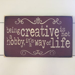 May include: A barn red wooden sign with cream text that reads "being creative is not a hobby, it's a way of life". The sign has decorative swirls at the top and bottom.